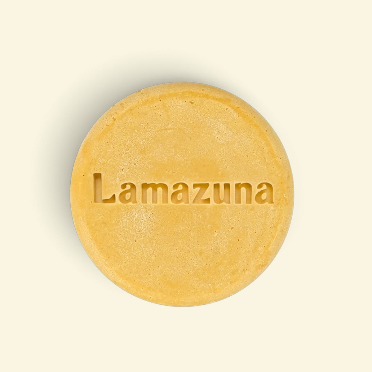 A round, textured yellow bar of soap with the brand name 'Lamazuna' embossed in the center, set against a solid cream-colored background.