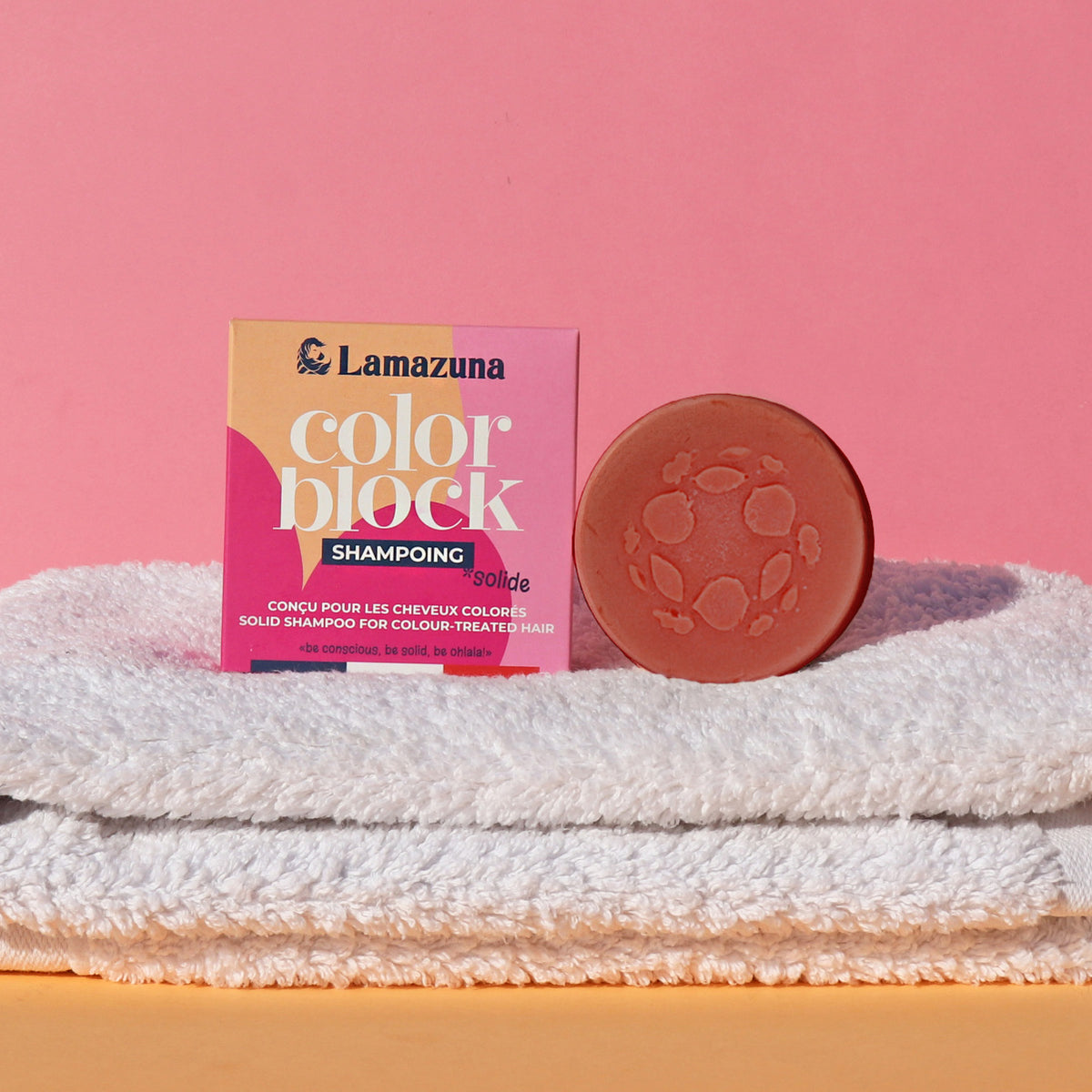 Solid shampoo bar and packaging on a white towel with pink background