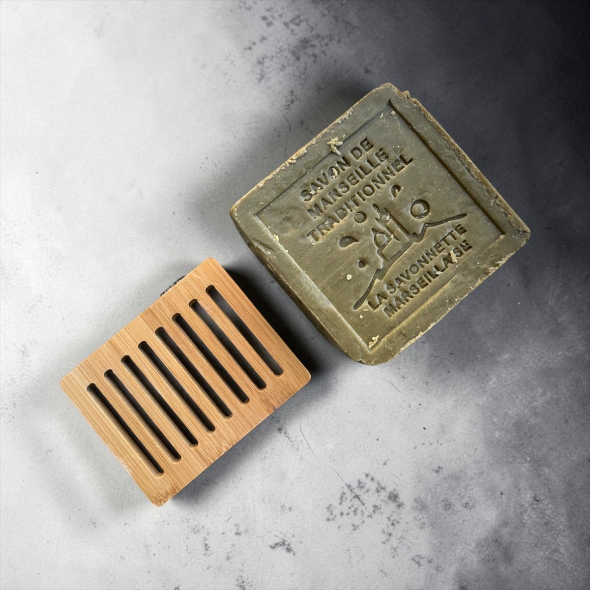 A rectangular bar of natural soap with a French embossed label reading 'Savon de Marseille Naturel' and 'La Savonnette Marseillaise' lies on a textured gray surface next to a small bamboo soap dish.