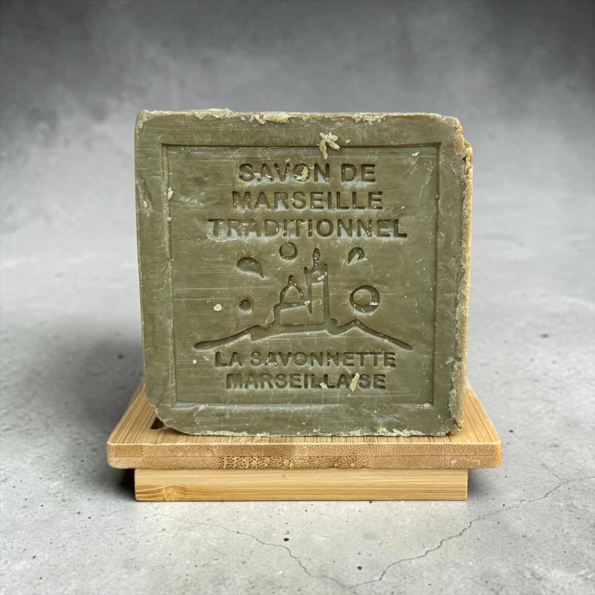 A square bar of traditional Marseille soap in an olive green color, resting on a small wooden display stand. The front of the soap is embossed with 'SAVON DE MARSEILLE TRADITIONNEL' and the brand 'LA SAVONNETTE MARSEILLAISE'.
