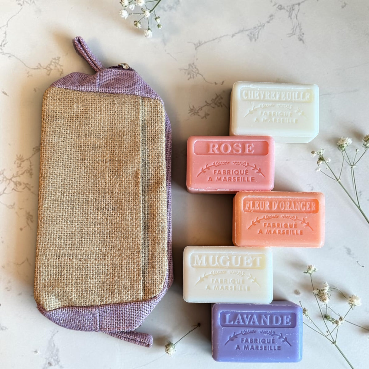 A lavender and beige woven pouch with a top loop, arranged on a light marble surface next to five rectangular bars of soap in white, pink, peach, cream, and lavender. Small white flowers are placed around the items.