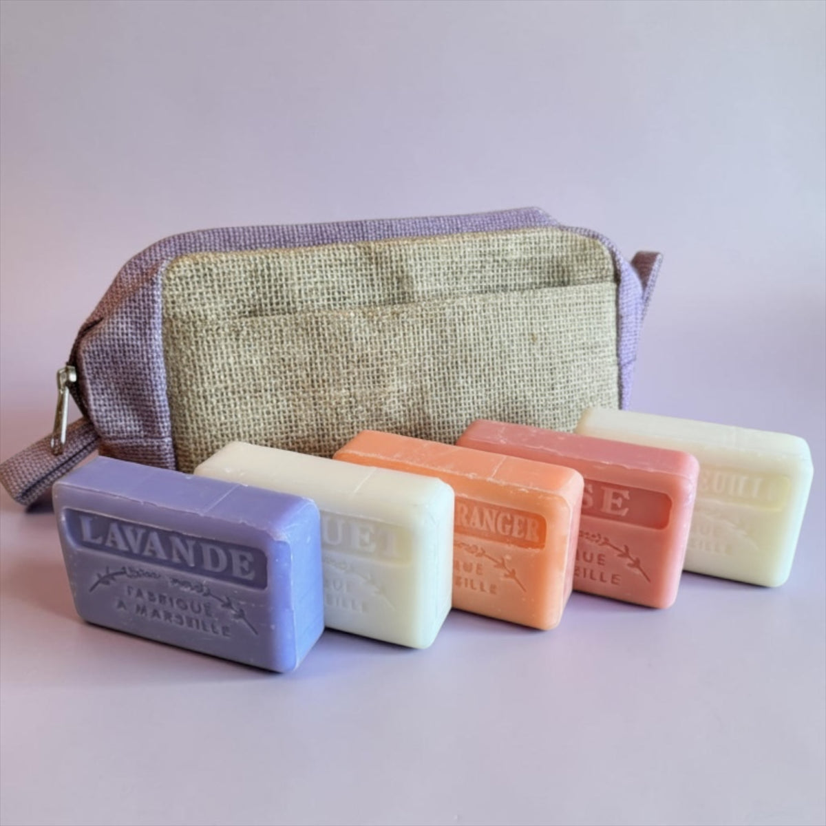 Five rectangular bars of handmade soap in lavender, cream, peach, and pinkish-orange are lined up in front of a small zippered pouch with a burlap-style front panel and lavender fabric trim. The soap bars are embossed with the brand name 'L'ANGE RICE'.