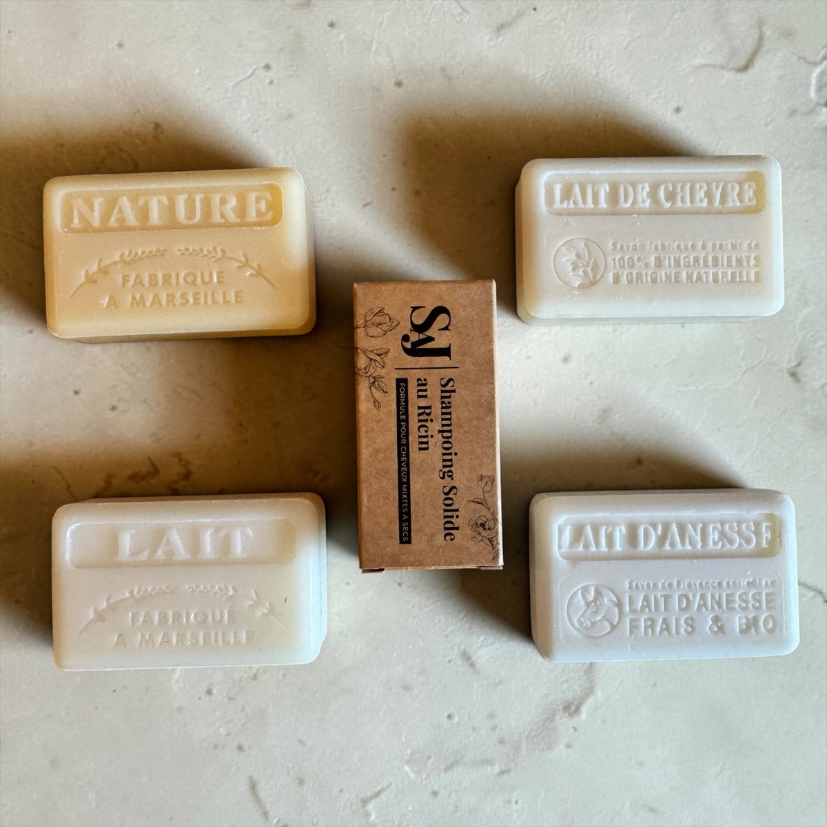 Four natural soaps and a central brown paper box arranged on a light stone surface. The soaps are labeled NATURE, LAIT DE CHEYRE, LAIT D'ANESSE, and LAIT DU MARISSILLE, with the center box reading Shampoie solide au Rich.