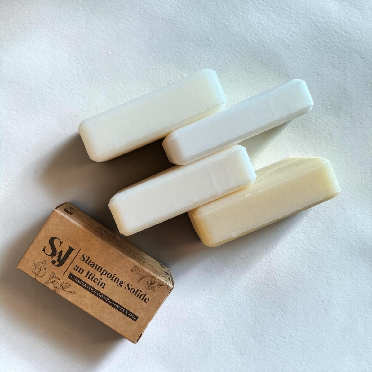 Four solid shampoo bars in off-white and cream shades arranged on a white surface next to a brown kraft paper box labeled 'SJ Shampooing Solide antichimica'.