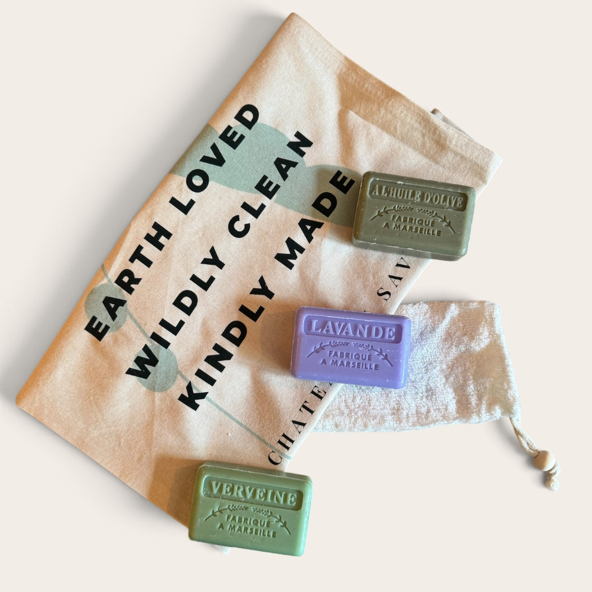 Three rectangular bars of soap in olive green, lavender purple, and sage green are arranged on a beige canvas drawstring bag with black text reading 'EARTH LOVED WILDLY CLEAN KINDLY MADE.' The soaps are embossed with their respective names and '100% ORGANIC COLDWEAR SOAP.