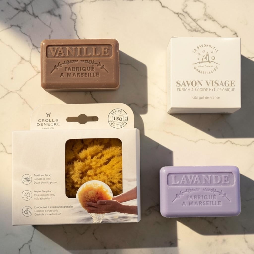 Four natural soap bars and their packaging arranged on a light marble surface. The set includes a brown bar labeled VANILLE, a white box labeled SAVON VISAGE, a white package with a yellow sponge, and a lavender bar labeled LAVANDE.
