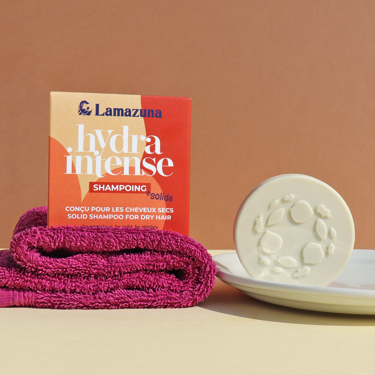 Lamazuna Hydra Intense Solid Shampoo for dry hair, featuring a red and beige cardboard box, a folded magenta towel, and a round white solid shampoo bar on a white plate against a warm beige background.