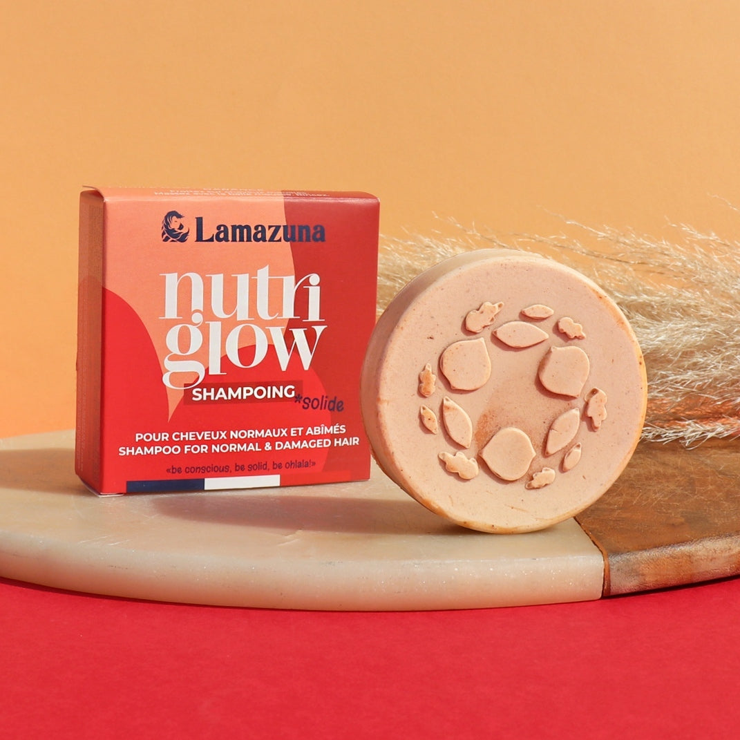 Lamazuna NutriGlow Solid Shampoo bar next to its red packaging box, styled on a wooden board with dried grass against a warm orange and red background.