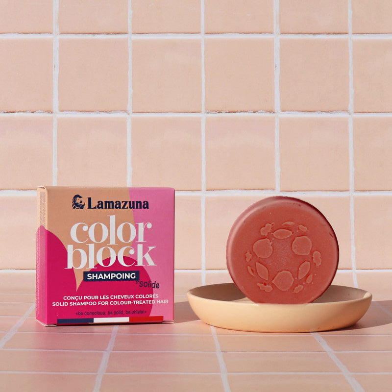 Lamazuna Color Block Shampoo solid bar shown next to its pink and beige packaging box, resting on a small beige dish against a tiled background.