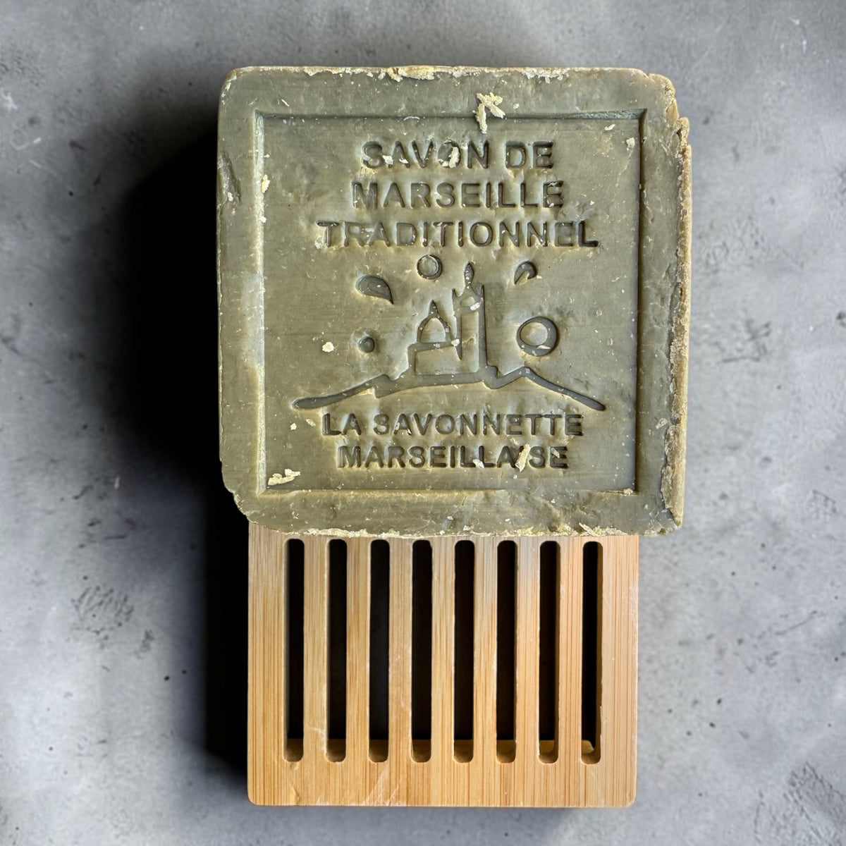 A rectangular bar of Marseille soap in a light olive-green color, embossed with 'SAVON DE MARSEILLE TRADITIONNEL' and 'LA SAVONNETTE MARSEILLAGE'. The soap is displayed on top of a bamboo matchstick-style holder against a textured gray concrete background.
