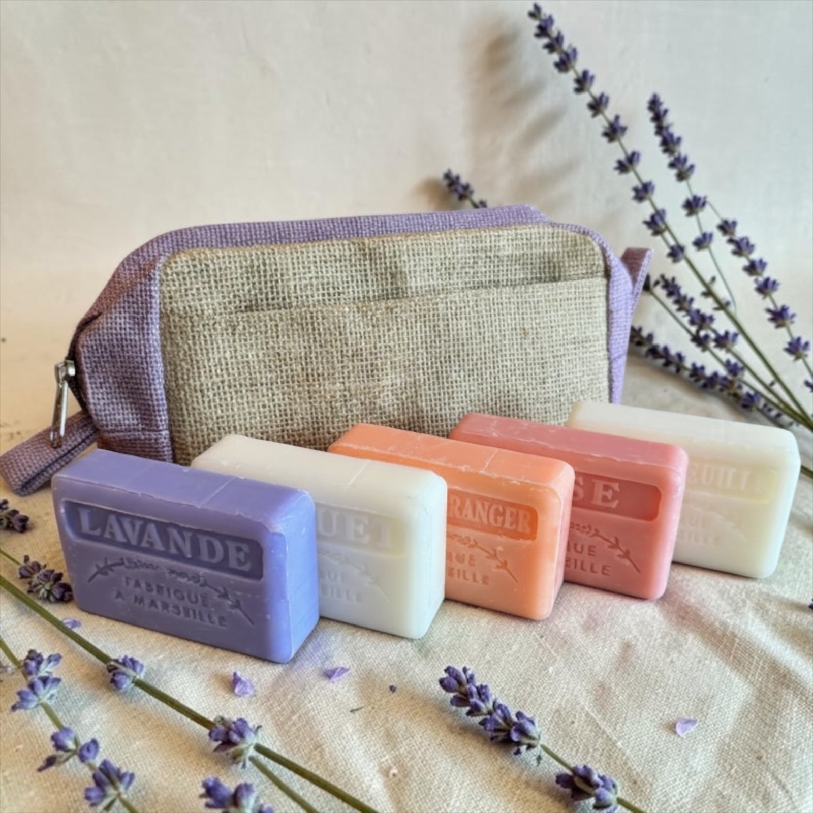 Four lavender soap bars in purple, white, peach, and cream are lined up in front of a lavender and burlap zip pouch on a beige fabric surface, surrounded by sprigs of dried lavender.