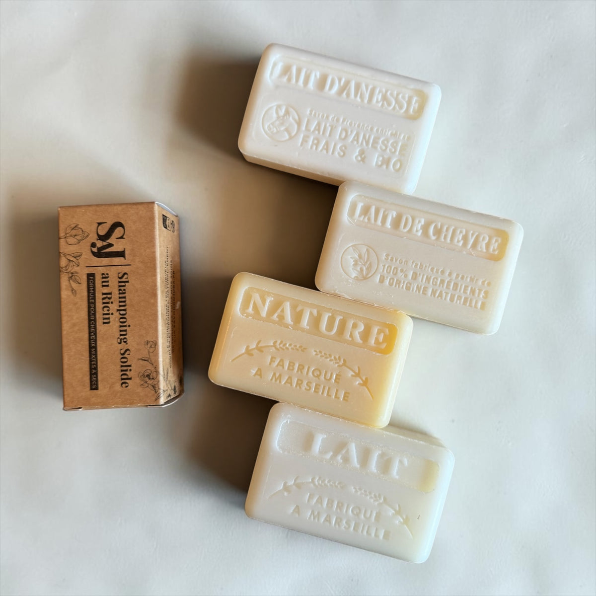 Four rectangular bars of Lait de Chyvre natural soap in off-white, cream, and pale yellow shades, arranged next to their brown cardboard packaging box on a light beige background.