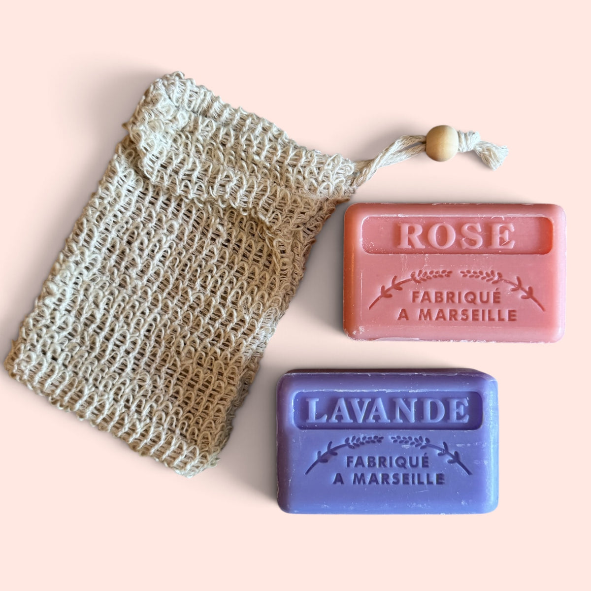 Two rectangular bars of Marseille soap in pink and purple, labeled ROSE and LAVANDE, displayed next to a natural beige cotton mesh soap bag on a solid pale pink background.