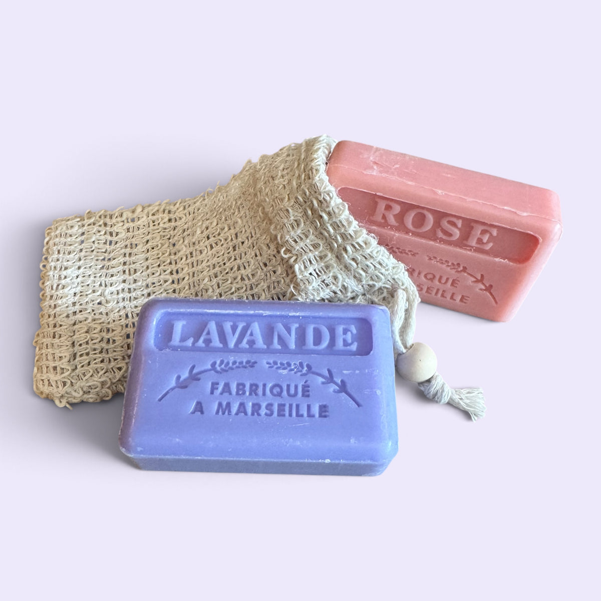 Two rectangular bars of Marseille soap, one lavender purple labeled 'LAVANDE' and one dusty pink labeled 'ROSE', arranged with a natural beige mesh soap bag on a light lavender background.