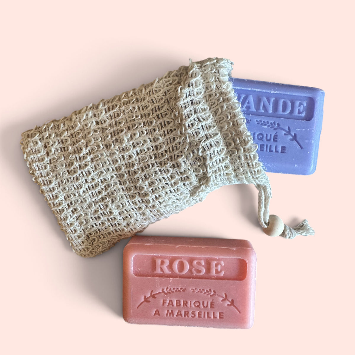 Two rectangular bar soaps, one pink labeled ROSE and one lavender-blue labeled VANDE, arranged with a knitted drawstring pouch on a pale peach background.