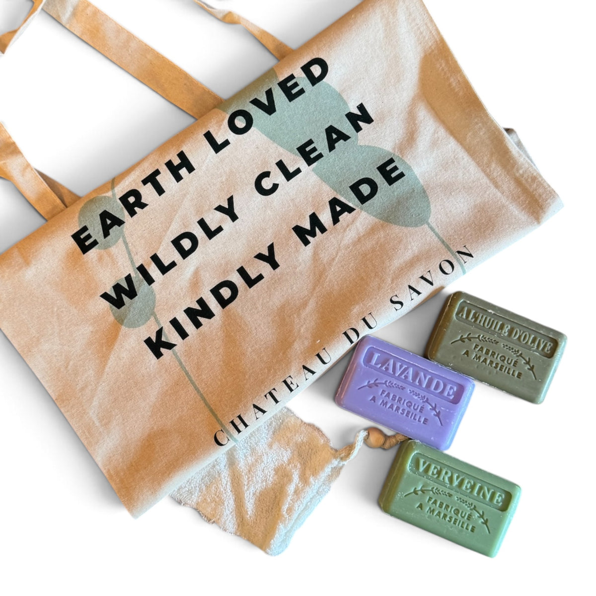 A natural canvas tote bag with black text reading 'EARTH LOVED WILDLY CLEAN KINDLY MADE' and 'CHATEAU DU SAVON,' shown with three rectangular bars of soap in lavender, olive green, and sage green on a white background.