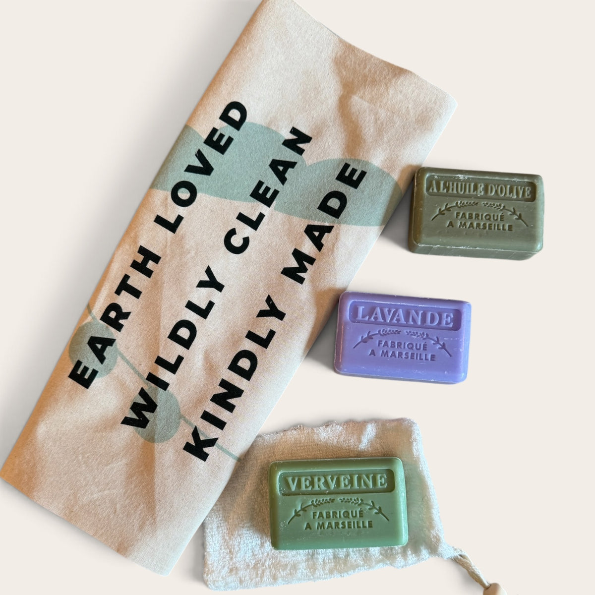 Three rectangular bars of handmade soap in green, purple, and mint green are arranged on a cream-colored fabric towel with black text reading 'EARTH LOVED WILDLY CLEAN KINDLY MADE.'.