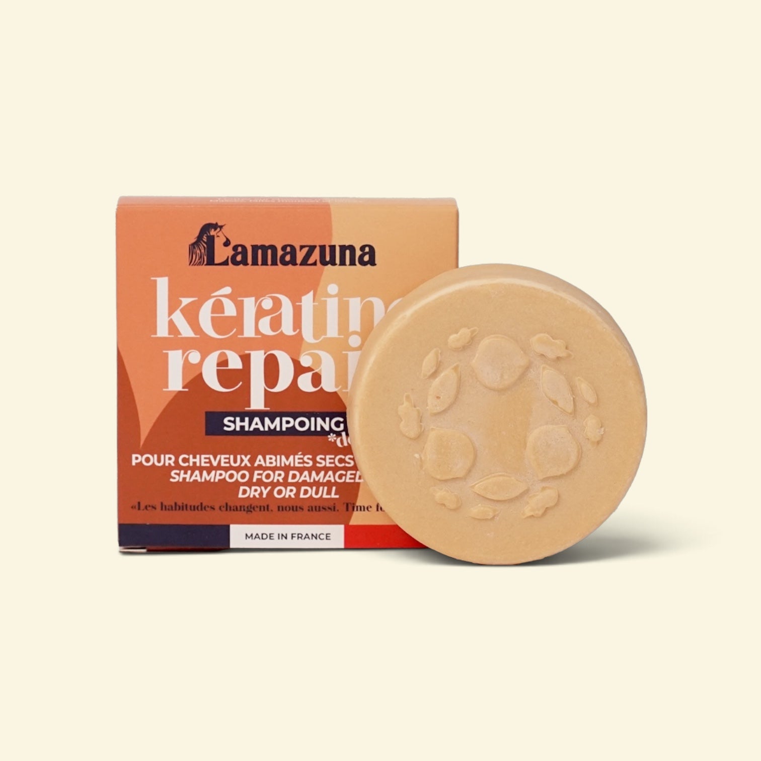 A round beige shampoo bar with an embossed floral pattern sits in front of its orange and white packaging box. The box is branded 'l'amazuna' and reads 'keratin repair shampoo'.