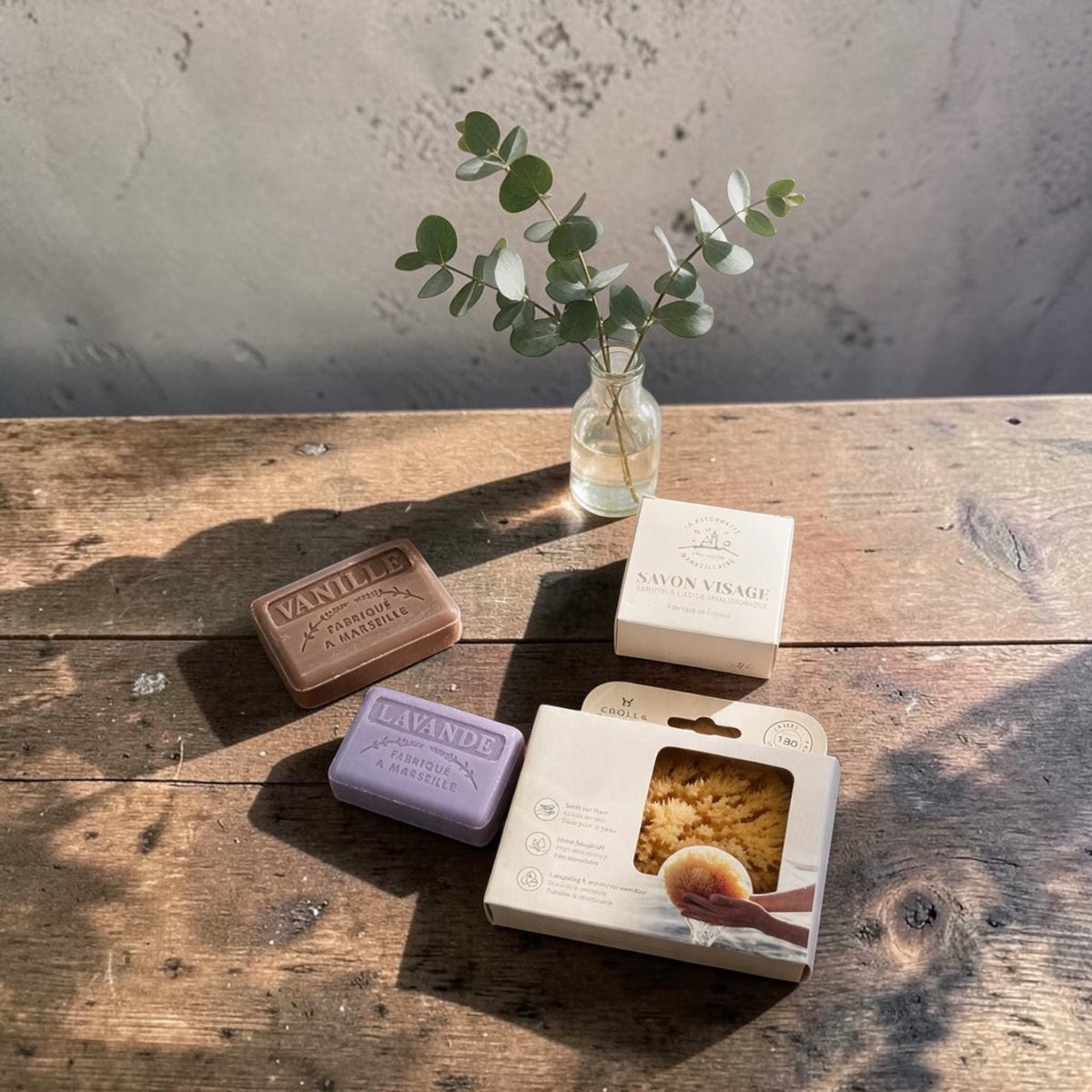 Three bars of soap and their boxes arranged on a rustic wooden table with a small glass vase of eucalyptus in the background. The soaps are in different colors: a brown bar labeled 'VANILLE', a lavender bar labeled 'LAVANDE', and white boxes labeled 'SAVON VISAGE'.